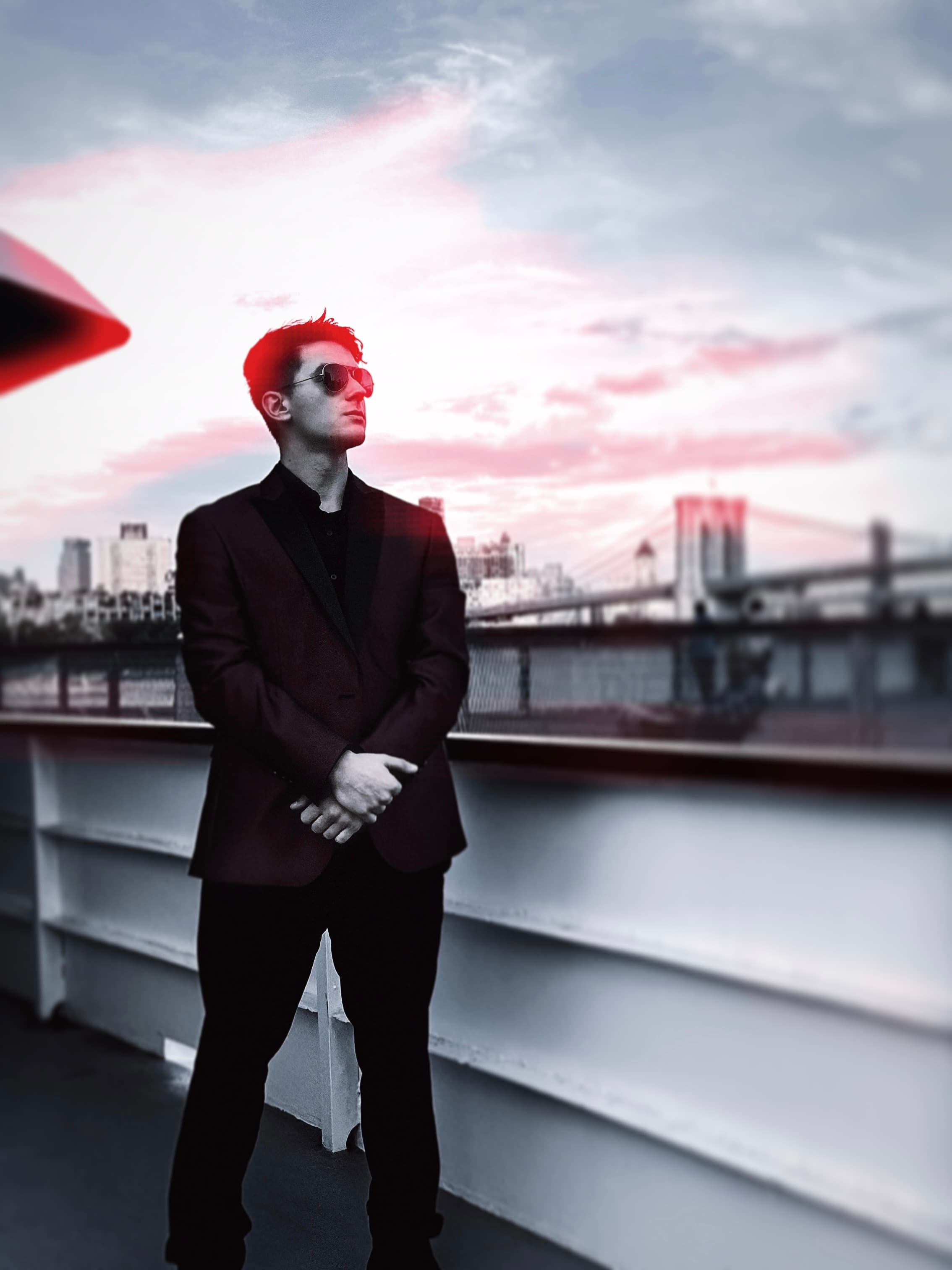 Anthony Savin in a dark suit and sunglasses on a boat deck, the Brooklyn Bridge behind him
